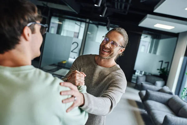 Excited and satisfied entrepreneur in eyeglasses shaking hands and touching shoulder while closing deal with business partner in modern lounge of coworking office, successful collaboration concept — Stock Photo
