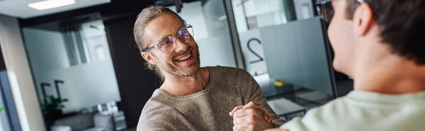 Overjoyed entrepreneur in stylish eyeglasses smiling and shaking hands with business partner in modern coworking environment, successful partnership concept, banner — Stock Photo