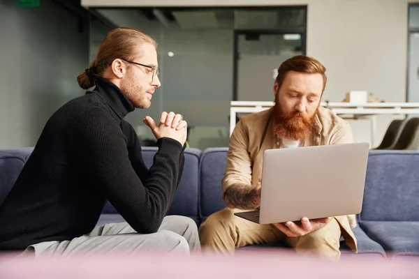 Thoughtful, bearded and tattooed entrepreneur working on startup project on laptop near serious businessman in black turtleneck and eyeglasses sitting in modern office, business partnership concept — Stock Photo
