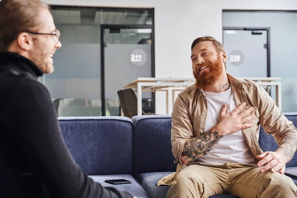 Благодарный, бородатый и татуированный предприниматель трогает сундук во время разговора с деловым партнером на диване в современной офисной среде, концепция делового сотрудничества — стоковое фото