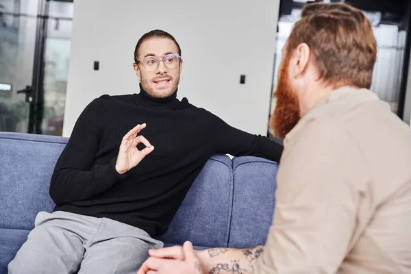 Businessman in eyeglasses and black turtleneck showing okay sign while planning startup project with bearded tattooed colleague in modern office, business collaboration concept — Stock Photo