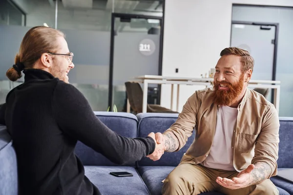 Delighted, bearded and tattooed entrepreneur sitting on couch near smartphone and shaking hands with business partner in black turtleneck in contemporary office, successful partnership concept — Stock Photo