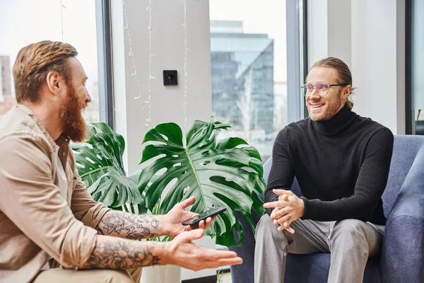 Bearded and tattooed entrepreneur holding smartphone and discussing business project with smiling colleague near green plant in contemporary office, business collaboration concept — Stock Photo