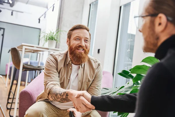 Overjoyed, tattooed and bearded entrepreneur in casual clothes confirming agreement and shaking hands with businessman on blurred foreground, successful partnership concept — Stock Photo