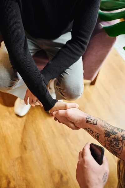 Top view of entrepreneur in black turtleneck shaking hands with tattooed businessman with mobile phone, cropped view, successful partnership concept — Stock Photo