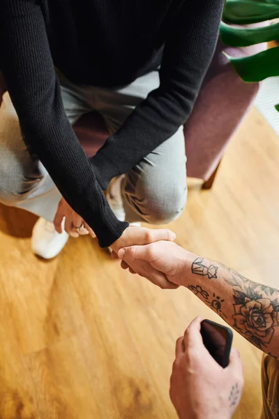 Vista dall'alto dell'imprenditore in dolcevita nero che stringe la mano con uomo d'affari tatuato con telefono cellulare, vista ritagliata, concetto di partnership di successo — Foto stock