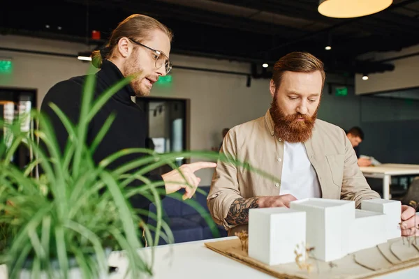 Creative architect pointing at building model and discussing startup project with bearded colleague near blurred plant in contemporary design studio, architecture and business concept — Stock Photo