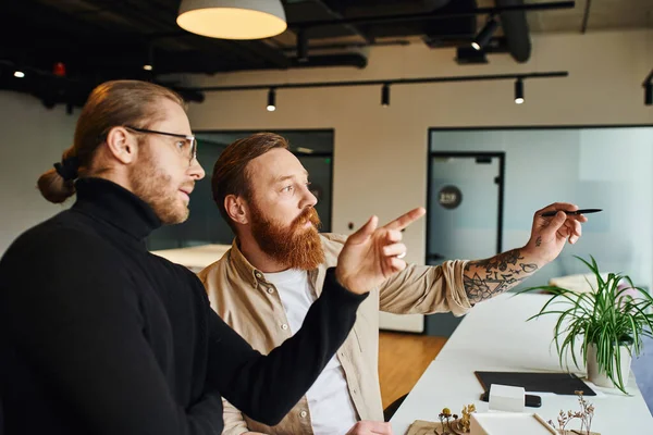 Creative and stylish architects looking away and pointing with finger and pen while working on startup project near building model in contemporary design studio, architecture and business concept — Stock Photo