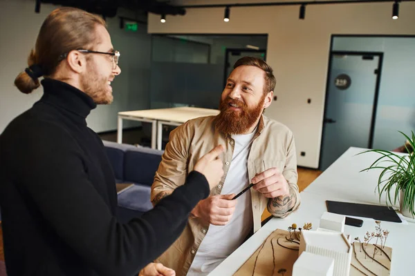 Smiling bearded architect holding pen and looking at happy colleague talking near building model while discussing startup project in modern design studio, architecture and business concept — Stock Photo