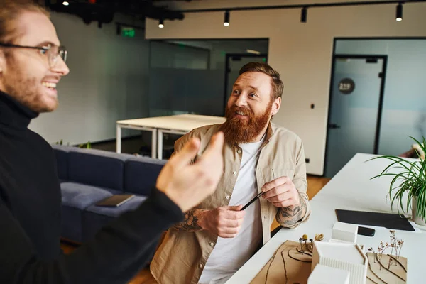 Inspired architect in black turtleneck discussing innovative ideas with cheerful, bearded and tattooed colleague near building model in modern design studio, architecture and business concept — Stock Photo