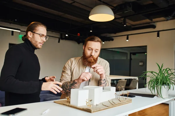 Thoughtful, tattooed and bearded architect looking at building model near colleague in black turtleneck while working on startup project in modern design studio, architecture and business concept — Stock Photo