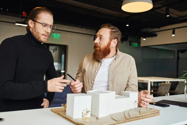 Worried bearded architect holding pen and talking to businessman in eyeglasses and black turtleneck while discussing startup near building model in design studio, architecture and business concept — Stock Photo