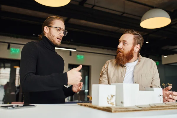 Attentive and bearded architect listening to colleague in black turtleneck and eyeglasses talking near building model in design studio, architecture and business concept — Stock Photo