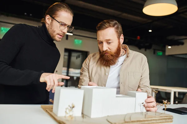 Seriöser Architekt mit Brille und schwarzem Rollkragen, der auf ein Baumodell zeigt und mit einem bärtigen Kollegen spricht, der in einem zeitgenössischen Designstudio, Architektur und Geschäftskonzept arbeitet — Stockfoto