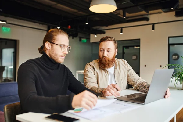 Businessman in eyeglasses and black turtleneck looking at papers with infographics near bearded, tattooed and thoughtful colleague sitting at laptop, working on startup project in modern office — Stock Photo
