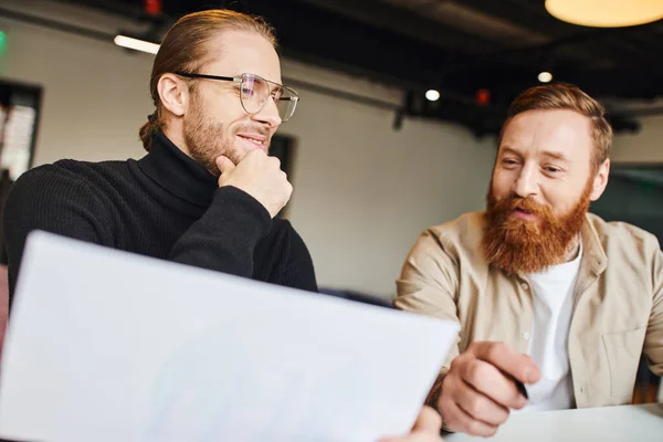 Complacido hombre de negocios con cuello alto negro y anteojos tocando la barbilla mientras mira el documento cerca de un colega barbudo feliz mientras trabaja en una nueva startup en la oficina de coworking - foto de stock