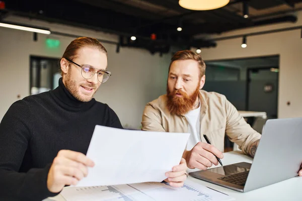 Smiling entrepreneur in black turtleneck and eyeglasses looking at paper near surprised bearded business partner, charts and laptop on work desk in contemporary office, startup planning concept — Stock Photo