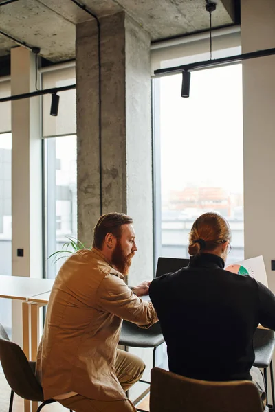 Вид ззаду бородатого бізнесмена з колегою в чорному черепашці, що сидить в сучасному офісному просторі з високотехнологічним інтер'єром і великими вікнами при роботі з інфографікою, концепцією стартап-планування — стокове фото