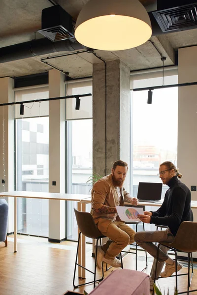 Hombre de negocios en gafas graduadas y cuello alto negro mirando cartas con un colega barbudo y tatuado mientras está sentado en el portátil con pantalla en blanco y planeando la puesta en marcha en un entorno de oficina moderno - foto de stock