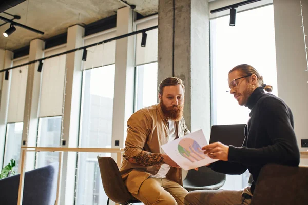 Stylish entrepreneurs sitting near laptop in office with high tech interior and looking at paper with charts, businessman in eyeglasses and black turtleneck with bearded and tattooed colleague — Stock Photo