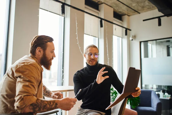 Inspired businessman in eyeglasses and black turtleneck showing folder with startup planning to bearded colleague in contemporary coworking office, productivity and collaboration concept — Stock Photo