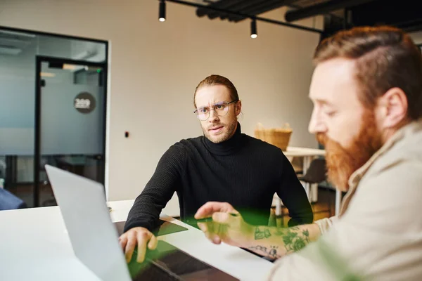 Bearded man pointing with pen at laptop near serious colleague in eyeglasses and black turtleneck, creative entrepreneurs working on startup project in modern office, blurred foreground — Stock Photo