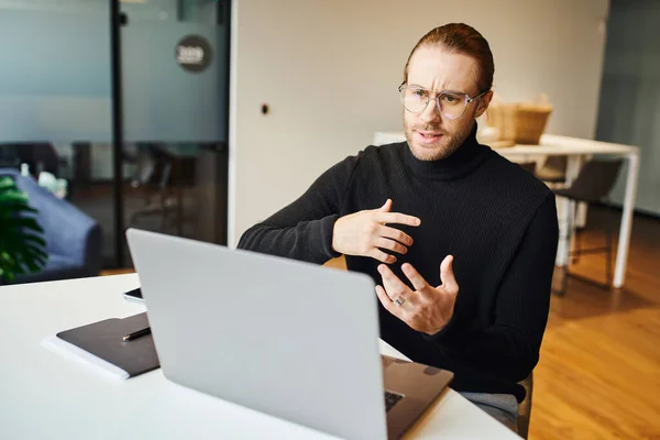 Businessman in black turtleneck and eyeglasses gesturing and talking during video conference on laptop while working in contemporary office space, business lifestyle concept — Stock Photo