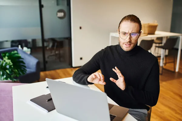 Serious entrepreneur in black turtleneck and eyeglasses gesturing and talking while having video call on computer in contemporary office space, business lifestyle concept — Stock Photo