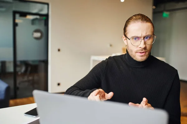 Серьезный предприниматель в черной водолазке и очках, имеющий видеозвонок на ноутбуке, говорящий и жестикулирующий в коворкинговой среде современного офиса, концепция бизнес-образа жизни — стоковое фото