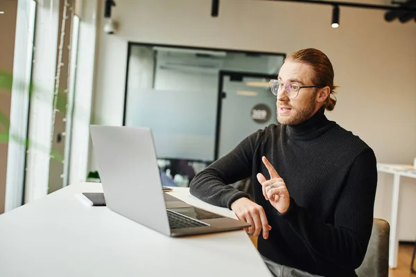 Hombre de negocios con estilo en cuello alto negro y gafas que muestran el gesto de atención y hablar durante el chat de vídeo en el ordenador portátil en el lugar de trabajo moderno en la oficina de coworking, concepto de estilo de vida de negocios - foto de stock