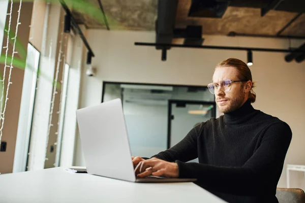 Homme d'affaires concentré à col roulé noir et lunettes de vue pensant près d'un ordinateur portable et travaillant sur un projet de démarrage dans un environnement de bureau moderne, concept de style de vie d'affaires — Photo de stock