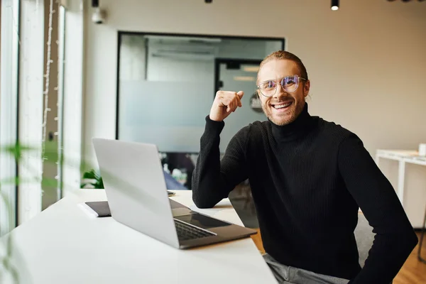 Happy entrepreneur in black turtleneck and eyeglasses sitting near laptop on work desk and looking at camera in coworking environment in modern office, business lifestyle concept — Stock Photo