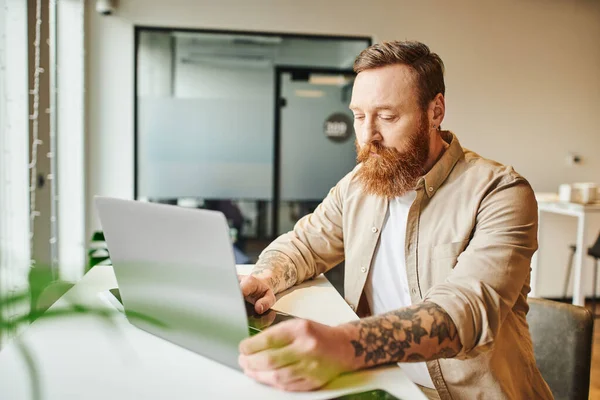 Серйозний бородатий і татуйований підприємець, який працює над стартап-проектом, сидячи біля ноутбука на робочому місці в сучасному коворкінгу, концепція ділового способу життя — стокове фото