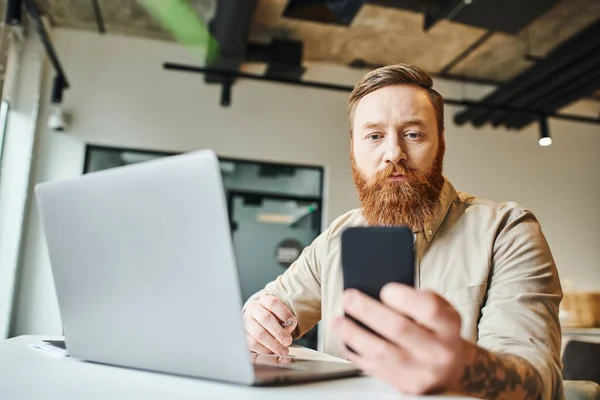 Серйозний бородатий бізнесмен сидить біля ноутбука і дивиться на мобільний телефон під час роботи над стартап-плануванням в сучасному офісному просторі з високотехнологічним інтер'єром, концепцією ділового способу життя — стокове фото