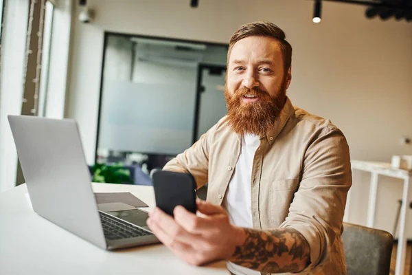 Happy tattooed and bearded entrepreneur holding smartphone and smiling at camera while sitting near laptop in contemporary coworking office, business lifestyle concept — Stock Photo