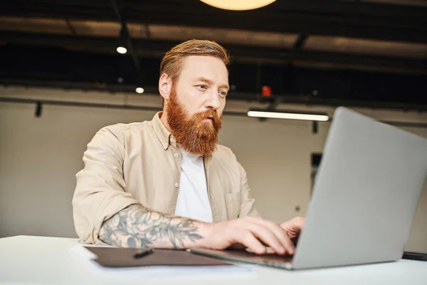 Целенаправленный татуированный и бородатый бизнесмен планирует стартап-проект во время работы на ноутбуке в коворкинг-среде в современном офисе, концепция бизнес-образа жизни — стоковое фото