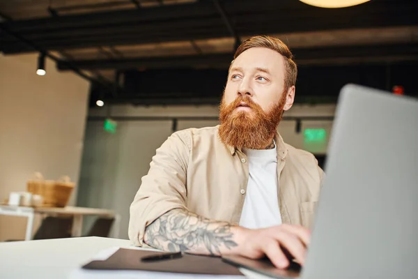 Вдумчивый, татуированный и бородатый предприниматель смотрит в сторону, сидя на рабочем месте, работая над проектом стартапа и печатая на ноутбуке в современном коворкинг пространстве, концепция бизнес-образа жизни — стоковое фото