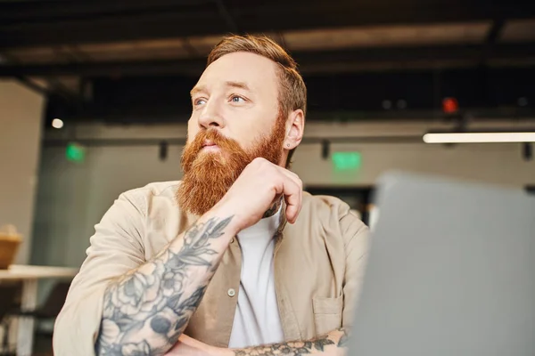 Portrait of pensive, tattooed and bearded entrepreneur sitting at workplace, looking away and thinking near laptop on blurred foreground in modern office, business lifestyle concept — Stock Photo