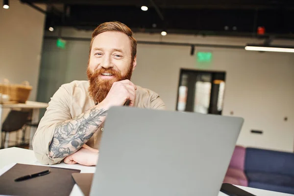 Весёлый, бородатый и татуированный предприниматель с сияющей улыбкой смотрит в камеру, сидя рядом с ноутбуком и работая над стартап-проектом, профессиональным фото, концепцией бизнес-образа жизни — стоковое фото