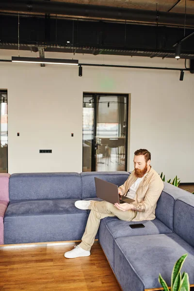 Полная длина татуированный и бородатый бизнесмен в стильной повседневной одежде работает на ноутбуке, сидя на удобном диване рядом со смартфоном в гостиной современного офиса, бизнес-концепция образа жизни — стоковое фото
