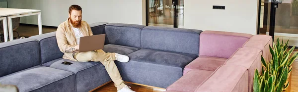 Pleine longueur d'entrepreneur élégant, barbu et tatoué assis sur un canapé confortable près du smartphone et travaillant sur ordinateur dans le salon du bureau contemporain, concept de style de vie d'affaires, bannière — Photo de stock