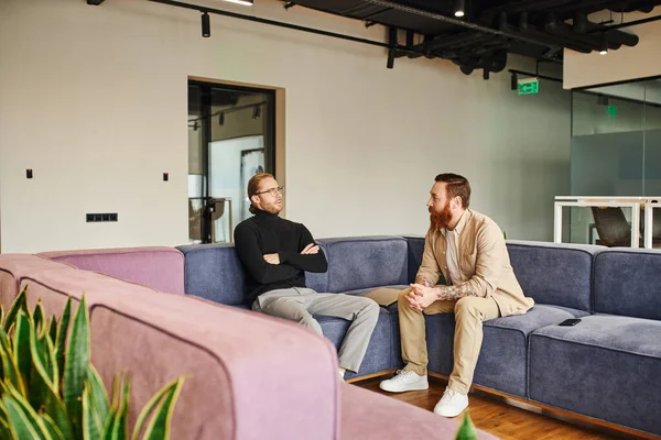 Homme barbu et tatoué parler à l'entrepreneur assis avec les bras croisés sur le canapé doux dans le salon de l'environnement de coworking contemporain, partenariat et concept de succès — Photo de stock