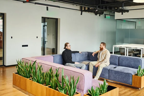 Серйозні бізнесмени в стильному повсякденному одязі, сидячи на м'якому дивані біля зелених рослин і обговорюючи стартап-проект в сучасному офісному лаунжі з високотехнологічним інтер'єром, партнерством і концепцією успіху — стокове фото