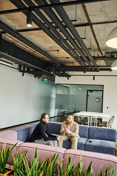 Bearded and tattooed businessman with colleague in black turtleneck and eyeglasses talking on soft couch in lounge of modern coworking office, partnership and success concept — Stock Photo
