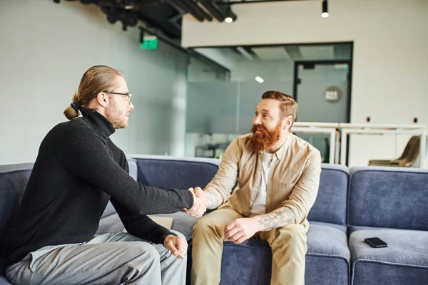 Happy bearded and tattooed man closing deal and shaking hands with entrepreneur in black turtleneck in lounge of contemporary coworking office, partnership and success concept — Stock Photo