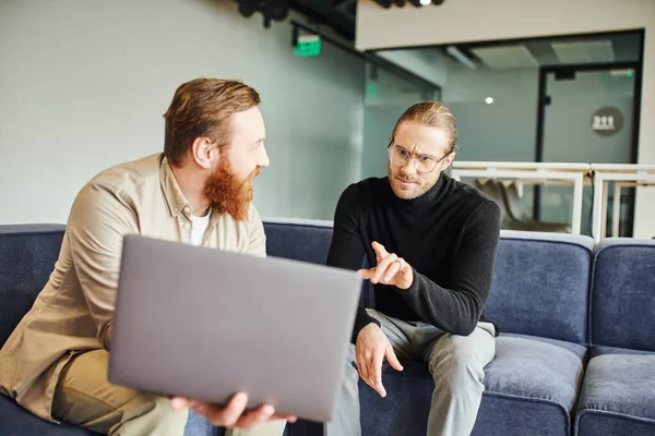 Displeased businessman in eyeglasses and black turtleneck pointing at laptop with startup project near bearded colleague while sitting in contemporary office, business partnership concept — Stock Photo