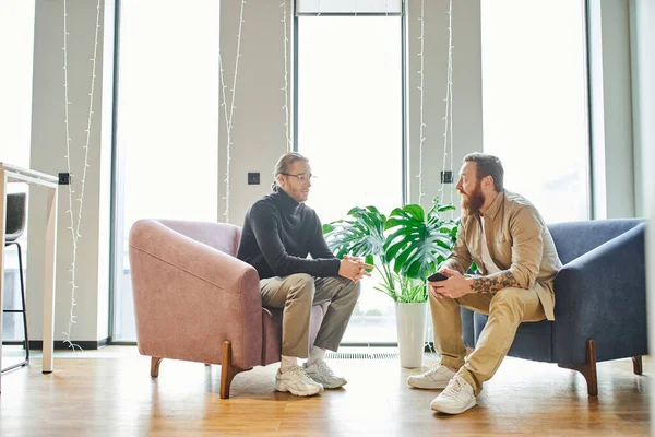 Full length of stylish business partners discussing possibility of startup projects in cozy armchairs near green plant and large windows in contemporary office, partnership and success concept — Stock Photo