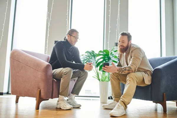 Bearded businessman showing mobile phone to colleague while talking in cozy armchairs in office lounge with large windows and high tech interior, partnership and success concept — Stock Photo