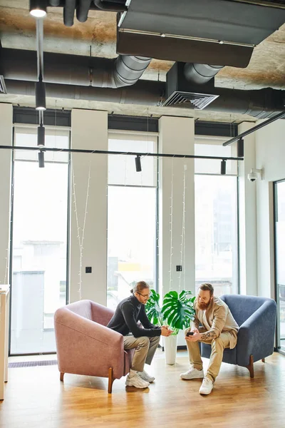 Full length of bearded man showing smartphone to colleague in black turtleneck while sitting in office lounge with cozy armchairs and large windows, partnership and success concept — Stock Photo