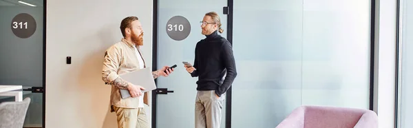 Side view of successful and stylish business partners with laptop and mobile phones standing in corridor of modern office and discussing startup project, productivity and cooperation concept, banner — Stock Photo